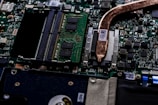 A close-up view of a computer motherboard showcasing various electronic components. The image displays integrated circuits, microchips, and a copper heat pipe. The green circuit board is densely packed with intricate pathways and connectors.