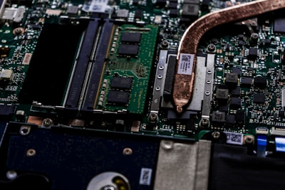 A close-up view of a computer motherboard showcasing various electronic components. The image displays integrated circuits, microchips, and a copper heat pipe. The green circuit board is densely packed with intricate pathways and connectors.