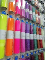 A display rack holding rolls of colorful vinyl wrapped in clear plastic, each roll is labeled and hung on hooks. The colors range from bright neon shades to metallic and glittery finishes.