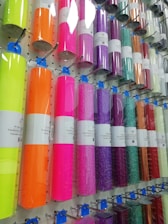 A display rack holding rolls of colorful vinyl wrapped in clear plastic, each roll is labeled and hung on hooks. The colors range from bright neon shades to metallic and glittery finishes.