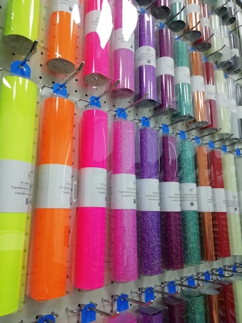A display rack holding rolls of colorful vinyl wrapped in clear plastic, each roll is labeled and hung on hooks. The colors range from bright neon shades to metallic and glittery finishes.
