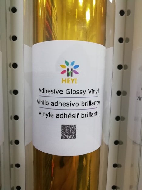 A roll of adhesive glossy vinyl is displayed with a white label featuring a colorful logo at the top. The label contains text in three languages: English, Spanish, and French, describing the product as adhesive glossy vinyl. A QR code is positioned below the text.