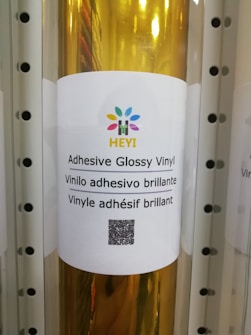 A roll of adhesive glossy vinyl is displayed with a white label featuring a colorful logo at the top. The label contains text in three languages: English, Spanish, and French, describing the product as adhesive glossy vinyl. A QR code is positioned below the text.