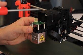 A hand is adjusting or assembling a component on a 3D printer. The component in focus appears to have a warning label with a laser symbol. The background includes various parts of the 3D printer such as rods and belts.