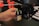 A hand is adjusting or assembling a component on a 3D printer. The component in focus appears to have a warning label with a laser symbol. The background includes various parts of the 3D printer such as rods and belts.