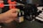 A hand is adjusting or assembling a component on a 3D printer. The component in focus appears to have a warning label with a laser symbol. The background includes various parts of the 3D printer such as rods and belts.