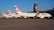 A line of Swiss airlines planes is parked on an airport tarmac. The aircraft have a distinct red and white tail design with a cross, and the word 'SWISS' is visible on the fuselage. In the background, there is an airport control tower and several lights.