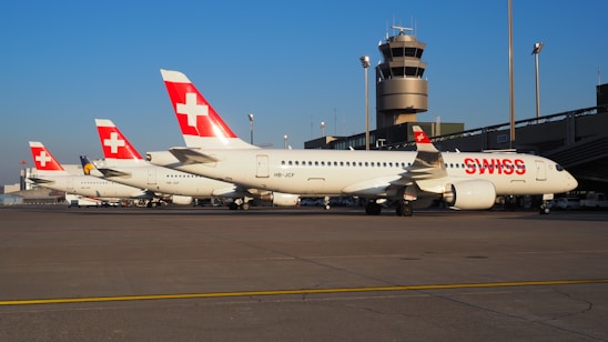 A line of Swiss airlines planes is parked on an airport tarmac. The aircraft have a distinct red and white tail design with a cross, and the word 'SWISS' is visible on the fuselage. In the background, there is an airport control tower and several lights.