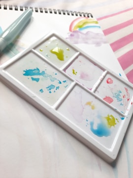 A watercolor palette with various colors, mostly green, pink, and blue, on a white tray. Next to it, a spiral-bound sketchbook with a painted rainbow and a water brush pen can be seen.