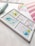 A watercolor palette with various colors, mostly green, pink, and blue, on a white tray. Next to it, a spiral-bound sketchbook with a painted rainbow and a water brush pen can be seen.
