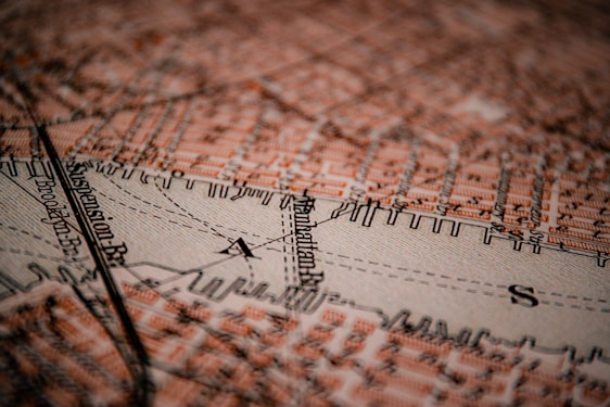 A detailed close-up of a vintage map featuring the Manhattan area. Streets and landmarks are marked in an orange-brown color, with bold black lines denoting paths and boundaries. The texture of the map is visible, providing an aged, tactile quality.
