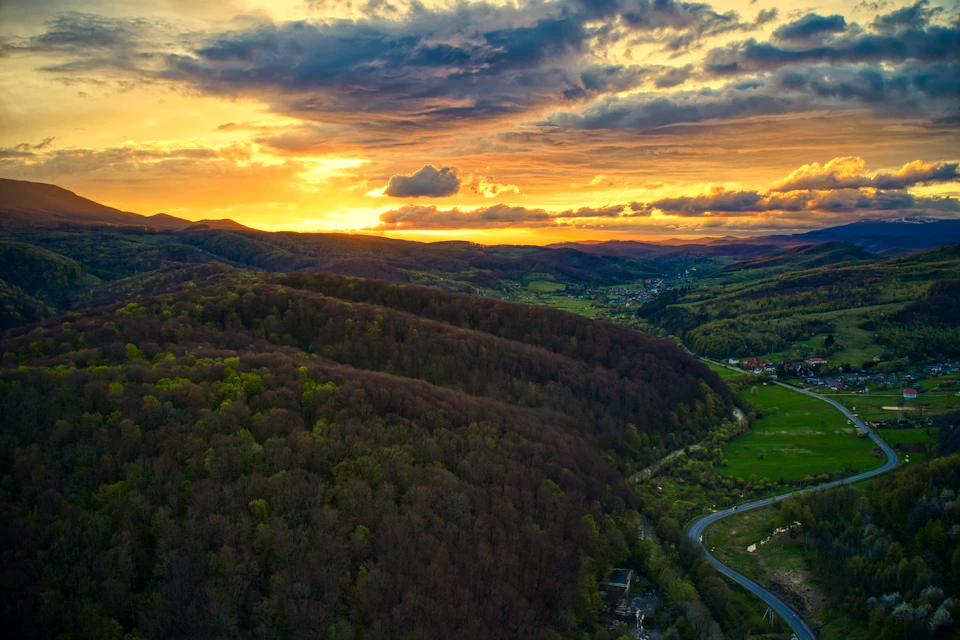 The image presents an aerial view of a valley nestled between two hills, with a winding road cutting through it. The valley is lush and green, dotted with trees that add depth to the scene. The sky above is painted in shades of orange and yellow, suggesting either sunrise or sunset. The perspective from above provides a comprehensive view of the landscape below, showcasing the natural beauty of the area.