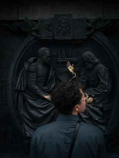 A person stands in front of a relief sculpture depicting two seated figures in historical attire, with golden hands emerging from the sculpture. Above them, smaller sculpted religious figures are visible.