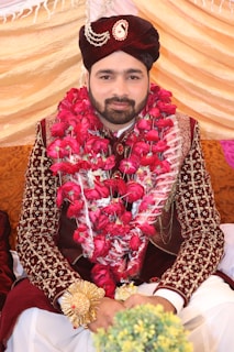 A man dressed in traditional attire sits adorned with a garland of vibrant red roses. He wears an intricately designed maroon velvet sherwani with detailed embroidery, and a matching turban embellished with jewels. The background consists of draped fabric in shades of gold and pink, indicating a celebratory or ceremonial setting.