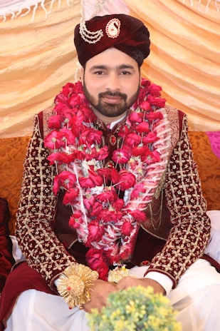 A man dressed in traditional attire sits adorned with a garland of vibrant red roses. He wears an intricately designed maroon velvet sherwani with detailed embroidery, and a matching turban embellished with jewels. The background consists of draped fabric in shades of gold and pink, indicating a celebratory or ceremonial setting.