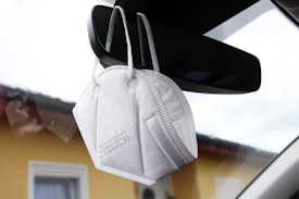A white protective face mask is hanging from a car's rearview mirror inside the vehicle. The background shows a blurred view of a yellow building with windows, seen through the car window.