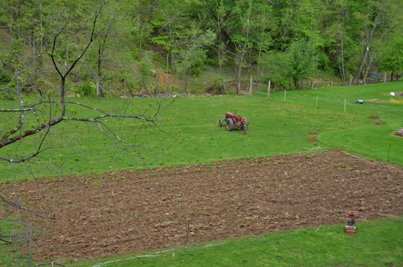 A lush, green landscape featuring a field in the foreground being tilled, while a red tractor continues plowing. The scene is surrounded by dense woods with various shades of green foliage. The ground is mostly made up of turned soil and patches of grass, creating a contrast between the earthy tones and the vibrant greens.