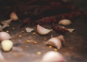 A close-up view of garlic cloves scattered on a dark surface, alongside dried red chili peppers and a few yellow seeds. The focus is on one garlic clove, with the background softly blurred, giving it an intimate and rustic feel.