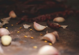 A close-up view of garlic cloves scattered on a dark surface, alongside dried red chili peppers and a few yellow seeds. The focus is on one garlic clove, with the background softly blurred, giving it an intimate and rustic feel.