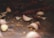 A close-up view of garlic cloves scattered on a dark surface, alongside dried red chili peppers and a few yellow seeds. The focus is on one garlic clove, with the background softly blurred, giving it an intimate and rustic feel.