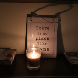 A small candle in a glass holder is lit, casting a warm glow on a hanging plaque with the inscription 'There is no place like home'. The plaque is placed against a wall, and its shadow is visible in the soft lighting. The setup is cozy and intimate, creating a sense of warmth and homeliness.