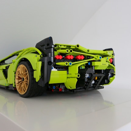 A detailed model car constructed from interlocking toy bricks is shown. The car features prominent lime green coloring with black and red accents and has gold-colored wheels. The intricate design includes visible components like gears and axles, showcasing a realistic back-end view of a sports car.