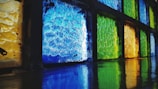 A series of textured glass blocks in various colors, including blue, green, and yellow, arranged in a grid pattern. The glass casts colorful reflections on the surface below, creating a vibrant and dynamic visual effect.