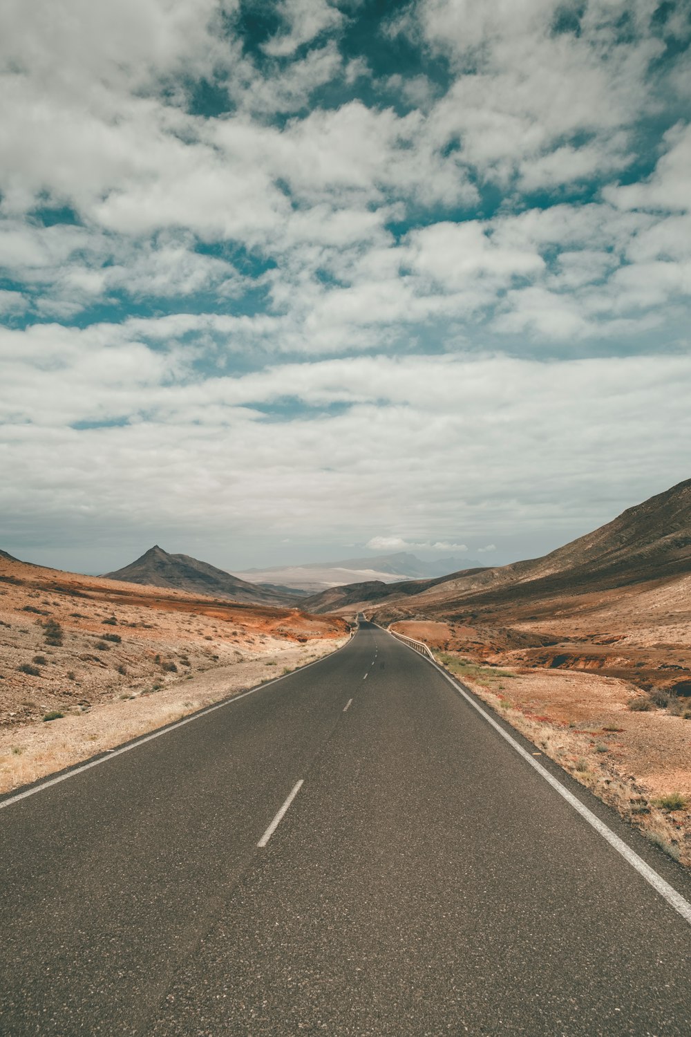 Carretera de asfalto negro entre montañas marrones bajo nubes blancas y cielo azul durante el día
