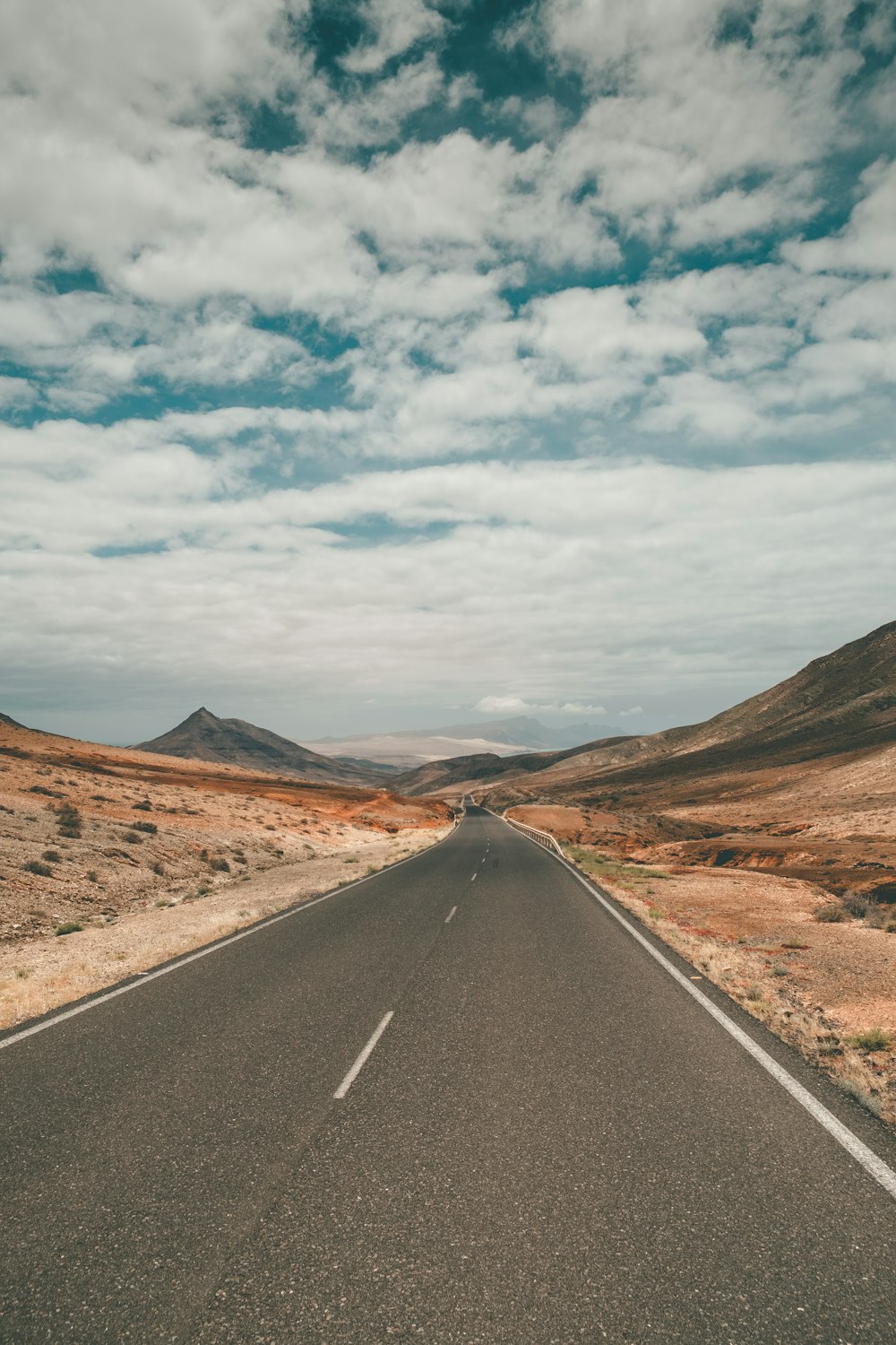 black asphalt road between brown mountains under white clouds and blue sky during daytime