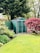 A well-manicured garden with a green metal storage shed in the background. The area is surrounded by lush trees and various plants, including a prominent red-leaved tree on the right.