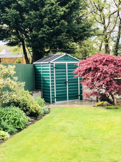 A well-manicured garden with a green metal storage shed in the background. The area is surrounded by lush trees and various plants, including a prominent red-leaved tree on the right.