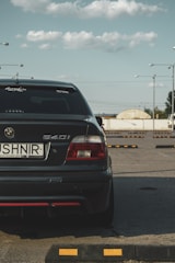 A dark-colored BMW 540i is prominently parked in an open parking area under a clear sky with scattered clouds. The car is seen from the rear, displaying a visible license plate with the name 'USHNIR' and decals on the windows. Surrounding the car are several parking blocks, with a few streetlights in the background and a distant view of a building.