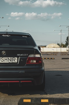 A dark-colored BMW 540i is prominently parked in an open parking area under a clear sky with scattered clouds. The car is seen from the rear, displaying a visible license plate with the name 'USHNIR' and decals on the windows. Surrounding the car are several parking blocks, with a few streetlights in the background and a distant view of a building.