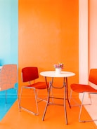 A brightly colored room features a modern setup with two red chairs and a white round table. The walls are painted orange and turquoise, creating a vibrant and cheerful atmosphere. A small pot of flowers is placed on the table, adding a touch of nature to the contemporary setting.