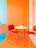 A brightly colored room features a modern setup with two red chairs and a white round table. The walls are painted orange and turquoise, creating a vibrant and cheerful atmosphere. A small pot of flowers is placed on the table, adding a touch of nature to the contemporary setting.