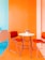 A brightly colored room features a modern setup with two red chairs and a white round table. The walls are painted orange and turquoise, creating a vibrant and cheerful atmosphere. A small pot of flowers is placed on the table, adding a touch of nature to the contemporary setting.