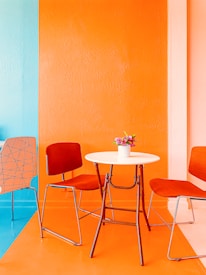 A brightly colored room features a modern setup with two red chairs and a white round table. The walls are painted orange and turquoise, creating a vibrant and cheerful atmosphere. A small pot of flowers is placed on the table, adding a touch of nature to the contemporary setting.