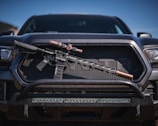 A tactical rifle with a scope is placed horizontally on the front grille of a dark-colored pickup truck. The rifle appears to have a black stock and barrel, with metallic components. The vehicle has a rugged appearance, with visible headlights and a sturdy bumper.