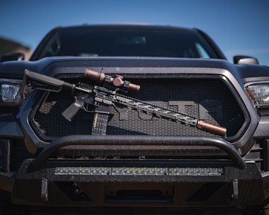 A tactical rifle with a scope is placed horizontally on the front grille of a dark-colored pickup truck. The rifle appears to have a black stock and barrel, with metallic components. The vehicle has a rugged appearance, with visible headlights and a sturdy bumper.