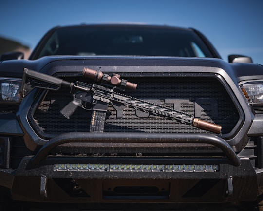 A tactical rifle with a scope is placed horizontally on the front grille of a dark-colored pickup truck. The rifle appears to have a black stock and barrel, with metallic components. The vehicle has a rugged appearance, with visible headlights and a sturdy bumper.