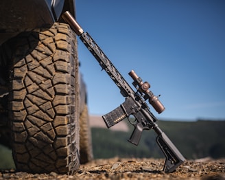 A rifle is leaning against the tire of a large off-road vehicle on rocky terrain. The weapon has a sleek design, equipped with a scope and a muzzle device, and is set in an outdoor setting with mountains and blue sky in the background.