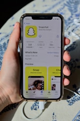 A hand holding an iPhone displaying the Snapchat app page on the App Store. The screen shows the app's logo, rating, age requirement, and previews. The phone is connected to a charger, resting on a patterned surface.