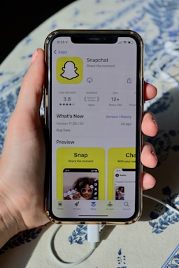 A hand holding an iPhone displaying the Snapchat app page on the App Store. The screen shows the app's logo, rating, age requirement, and previews. The phone is connected to a charger, resting on a patterned surface.