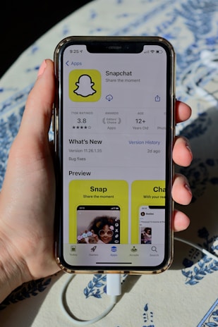 A hand holding an iPhone displaying the Snapchat app page on the App Store. The screen shows the app's logo, rating, age requirement, and previews. The phone is connected to a charger, resting on a patterned surface.