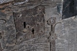 A close-up view of an old, rustic lock mounted on a weathered wooden surface. The lock and its surrounding metalwork exhibit signs of rust and age, with intricate, floral patterns carved into the metal. The wood shows worn and rough textures, enhancing the antique and historic character.