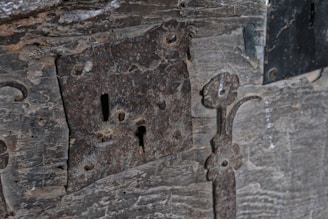 A close-up view of an old, rustic lock mounted on a weathered wooden surface. The lock and its surrounding metalwork exhibit signs of rust and age, with intricate, floral patterns carved into the metal. The wood shows worn and rough textures, enhancing the antique and historic character.