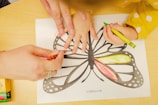 A child's hand is being guided by an adult hand to color in a butterfly drawing on paper using crayons. The butterfly drawing is being filled with different colors such as red and yellow. The overall setting appears to be a creative or educational activity.