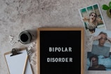 A letter board displays the words 'BIPOLAR DISORDER' surrounded by scattered letters. Nearby, there is a clipboard with a blank sheet of paper and a pencil, accompanied by a cup of black coffee. Three photographs show different facial expressions, and a small potted plant is visible. The surface is textured in a neutral tone.