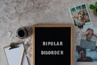 A letter board displays the words 'BIPOLAR DISORDER' surrounded by scattered letters. Nearby, there is a clipboard with a blank sheet of paper and a pencil, accompanied by a cup of black coffee. Three photographs show different facial expressions, and a small potted plant is visible. The surface is textured in a neutral tone.