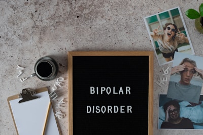 A letter board displays the words 'BIPOLAR DISORDER' surrounded by scattered letters. Nearby, there is a clipboard with a blank sheet of paper and a pencil, accompanied by a cup of black coffee. Three photographs show different facial expressions, and a small potted plant is visible. The surface is textured in a neutral tone.